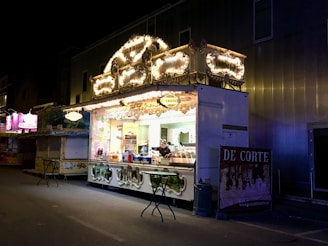 A brightly lit food stall adorned with decorative lights stands in contrast to the dimly lit surroundings. The stall's facade is ornately decorated with a vintage design and signage promoting fries. The setup includes folding tables outside, and a few people are visible inside, busy with preparations. Nearby, other closed stalls line the street.