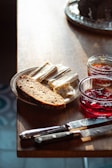 On a wooden table, there is a plate with slices of soft cheese and a piece of bread. Two glass jars contain different types of jam nearby. A couple of knives rest beside the plate, and natural lighting casts gentle shadows across the surface.