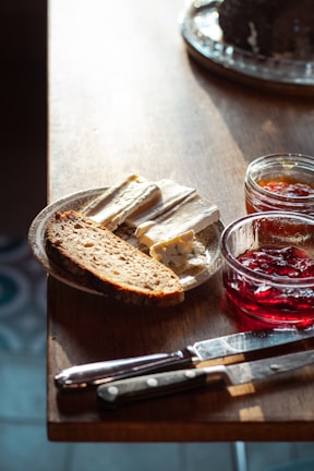 On a wooden table, there is a plate with slices of soft cheese and a piece of bread. Two glass jars contain different types of jam nearby. A couple of knives rest beside the plate, and natural lighting casts gentle shadows across the surface.