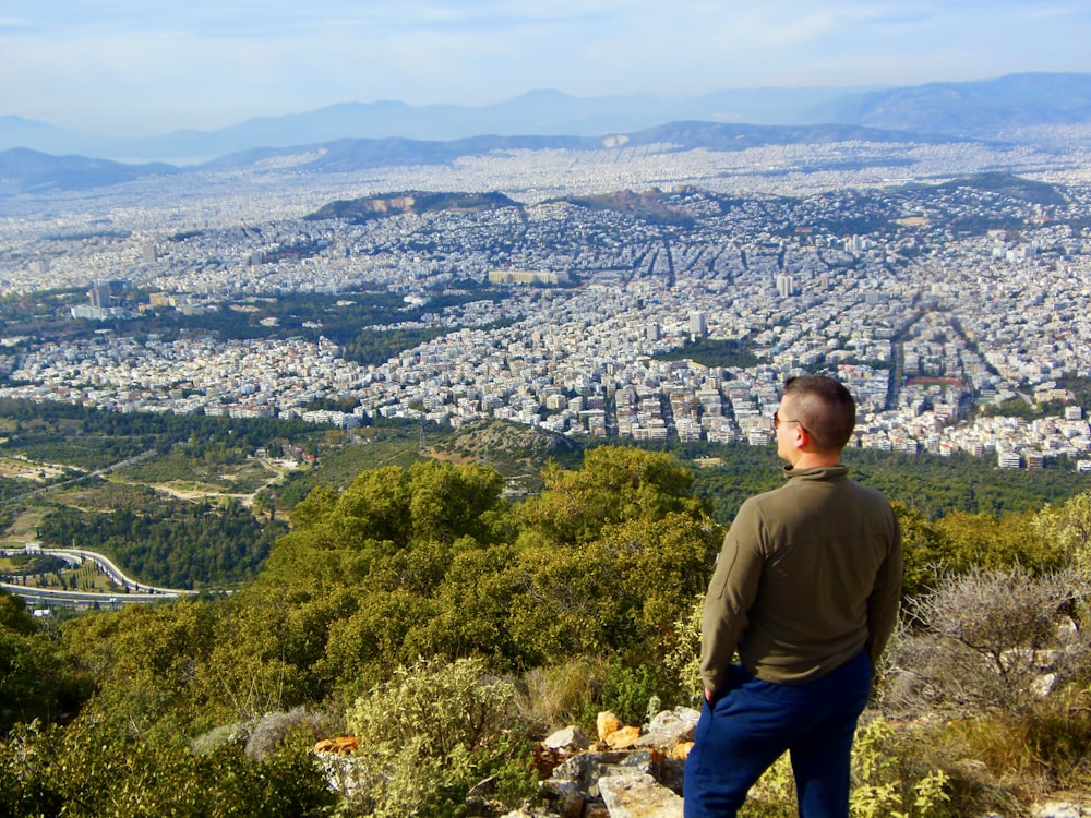 Mann im grünen Langarmhemd steht auf dem Gipfel des Berges und schaut tagsüber auf die Stadt