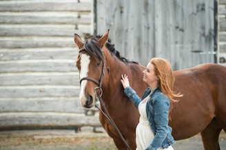 A person with long hair and wearing a denim jacket is gently petting a brown horse. The setting is outdoors, with a rustic wooden structure in the background.