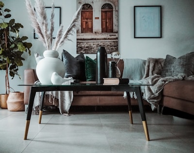 A modern living room features a glass-top coffee table with a white vase filled with tall dried pampas grass, decorative books, and a black bottle. The background includes a brown sofa adorned with a gray throw and velvet cushions in green and gray hues. Wall art consisting of a photograph and framed pictures is displayed above the sofa. A small white ceramic bird and an hourglass add to the décor. A potted plant with green leaves is situated in a woven basket to the left, complementing the stylish and cozy atmosphere.