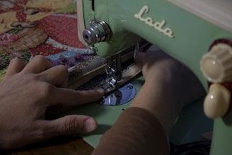 A person uses a vintage green sewing machine to stitch fabric with a colorful, patterned cloth. The focus is on the hands and the sewing process.