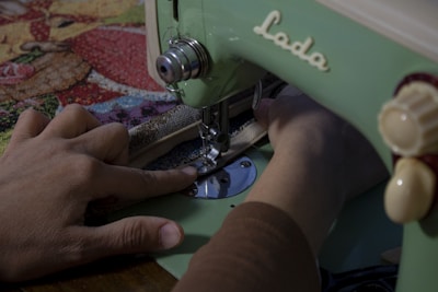 A person uses a vintage green sewing machine to stitch fabric with a colorful, patterned cloth. The focus is on the hands and the sewing process.