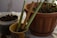A collection of gardening tools rests in a small ceramic pot beside a larger plant pot filled with soil and green stems. A plastic container with soil is placed nearby. The scene suggests an indoor gardening setup with a focus on planting or potting activities.