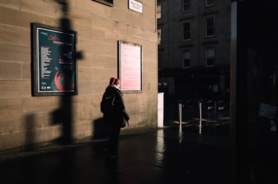 A person wearing a backpack stands on a dimly lit sidewalk next to a building with two large posters. The setting is urban, with shadows cast across the scene, and the ground appears to be wet, reflecting some light.