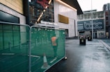 A person in an orange safety vest stands behind a green fence barrier near the entrance to Glasgow Queen Street. The street appears wet, possibly from rain. A construction cone is placed in front of the fence. There is a modern building facade on the left and a large sign on the building to the right saying 'bloc.' with more text that isn't fully visible.