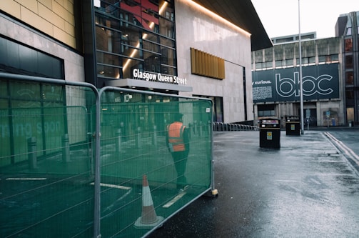 A person in an orange safety vest stands behind a green fence barrier near the entrance to Glasgow Queen Street. The street appears wet, possibly from rain. A construction cone is placed in front of the fence. There is a modern building facade on the left and a large sign on the building to the right saying 'bloc.' with more text that isn't fully visible.