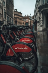 A row of parked rental bicycles with red coverings displaying a Santander logo, set on a wet street in an urban environment with historic architecture in the background. The scene is captured on a rainy day, evident from the glistening wet pavement.