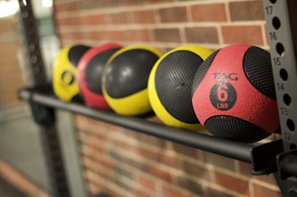 A row of four medicine balls neatly placed on a metallic rack against a brick wall. The balls are colored in yellow and red with black textured sections. Each ball is marked with weight measurements indicating 6 pounds on the red ball.