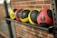 A row of four medicine balls neatly placed on a metallic rack against a brick wall. The balls are colored in yellow and red with black textured sections. Each ball is marked with weight measurements indicating 6 pounds on the red ball.