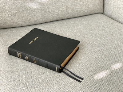 A hardcover book titled 'Holy Bible' rests on a light gray textured fabric surface. Black ribbons protrude from the book's pages, indicating it is often read and marked. The book's cover is dark green with gold lettering.
