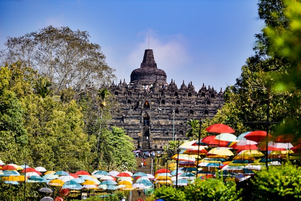 An ancient temple structure surrounded by lush greenery and colorful umbrellas neatly lining a pathway. The temple features intricate stone carvings and a prominent central dome, set against a clear blue sky.