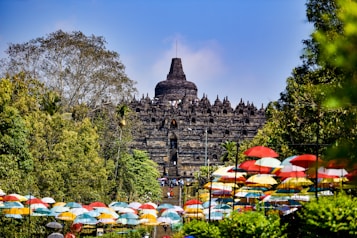 An ancient temple structure surrounded by lush greenery and colorful umbrellas neatly lining a pathway. The temple features intricate stone carvings and a prominent central dome, set against a clear blue sky.