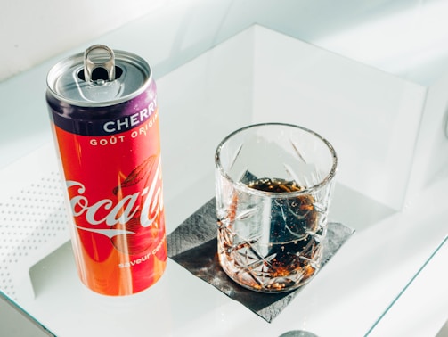 A can of cherry-flavored Coca-Cola and a glass filled with a dark carbonated beverage are placed on a reflective glass surface. The can has a vibrant red and purple color with branding visible, while the clear glass has a textured, decorative pattern and is placed on a black napkin.