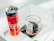 A can of cherry-flavored Coca-Cola and a glass filled with a dark carbonated beverage are placed on a reflective glass surface. The can has a vibrant red and purple color with branding visible, while the clear glass has a textured, decorative pattern and is placed on a black napkin.