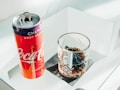 A can of cherry-flavored Coca-Cola and a glass filled with a dark carbonated beverage are placed on a reflective glass surface. The can has a vibrant red and purple color with branding visible, while the clear glass has a textured, decorative pattern and is placed on a black napkin.