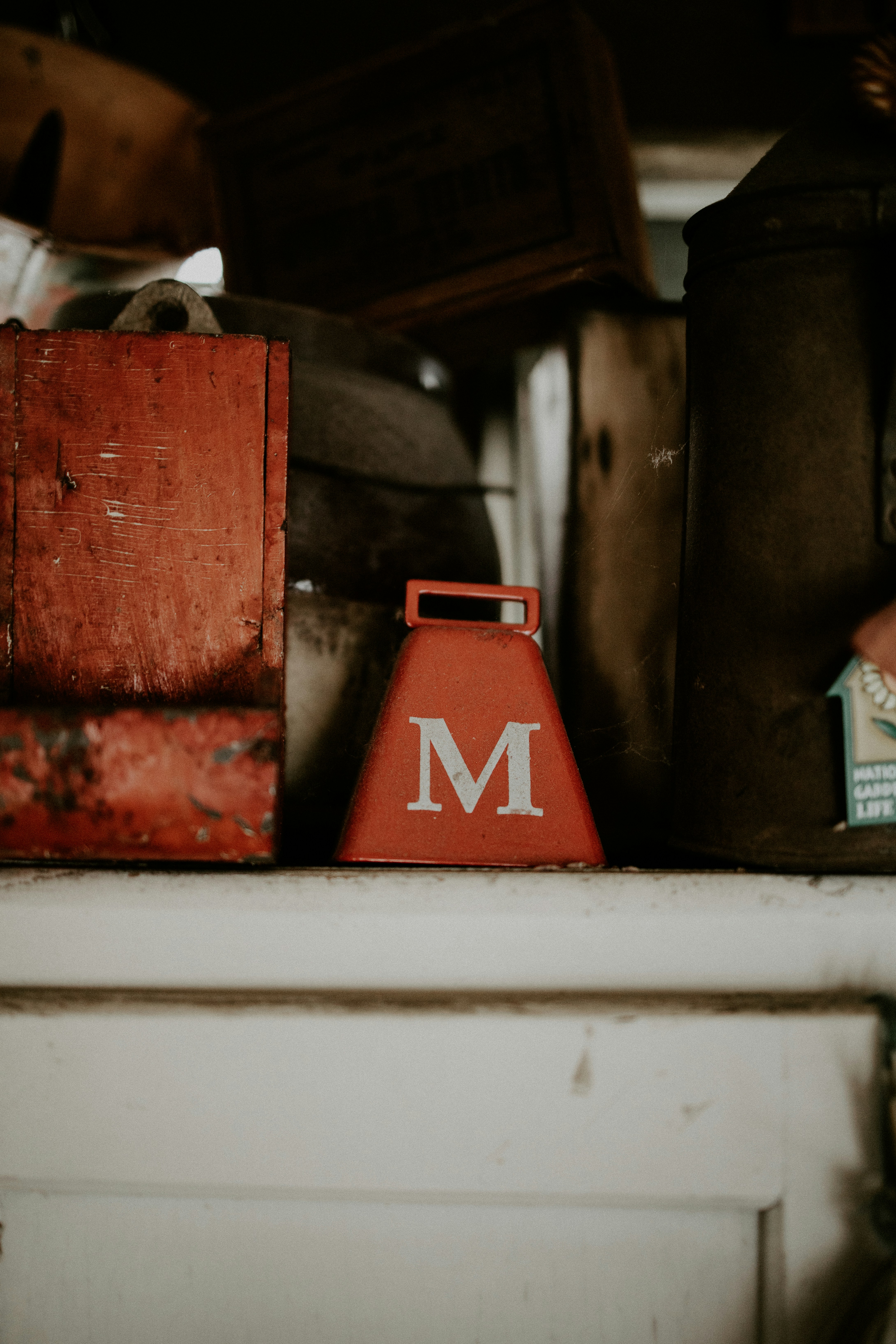 Red M-marked metal tool on a dusty shelf, surrounded by weathered containers and dark shadows.