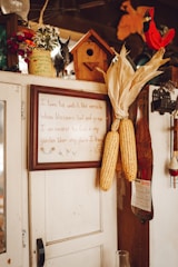 A rustic interior scene featuring a wooden cabinet adorned with various items. Corn ears are hung on the side of the cabinet. A framed quote is displayed prominently. A small birdhouse and decorative items like a calendar, a red cardinal figurine, and flower pots are placed on top.