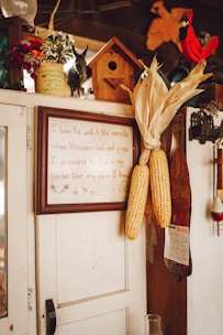 A rustic interior scene featuring a wooden cabinet adorned with various items. Corn ears are hung on the side of the cabinet. A framed quote is displayed prominently. A small birdhouse and decorative items like a calendar, a red cardinal figurine, and flower pots are placed on top.