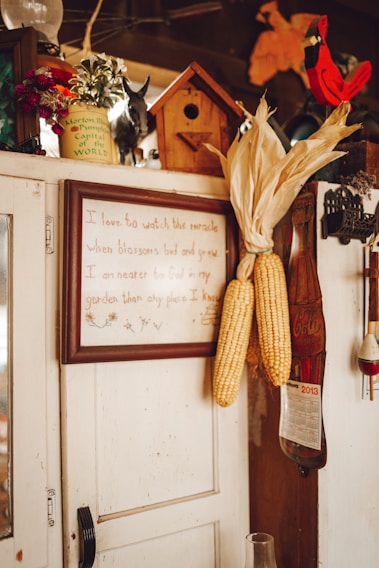 A rustic interior scene featuring a wooden cabinet adorned with various items. Corn ears are hung on the side of the cabinet. A framed quote is displayed prominently. A small birdhouse and decorative items like a calendar, a red cardinal figurine, and flower pots are placed on top.