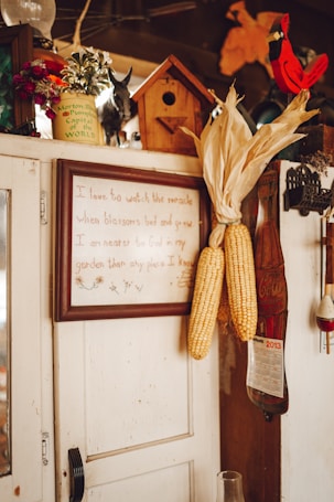 A rustic interior scene featuring a wooden cabinet adorned with various items. Corn ears are hung on the side of the cabinet. A framed quote is displayed prominently. A small birdhouse and decorative items like a calendar, a red cardinal figurine, and flower pots are placed on top.