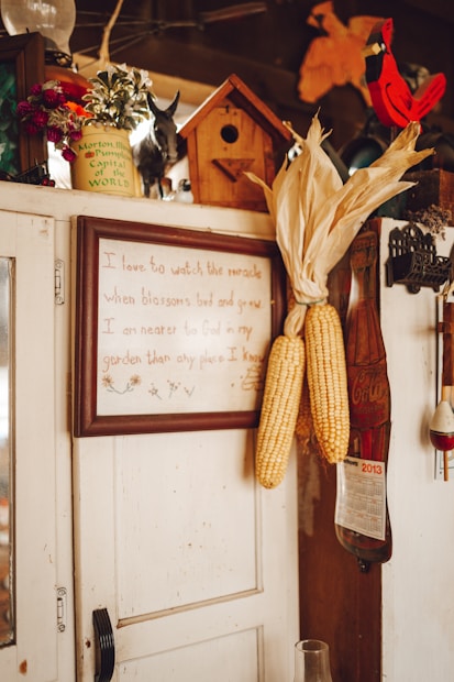 A rustic interior scene featuring a wooden cabinet adorned with various items. Corn ears are hung on the side of the cabinet. A framed quote is displayed prominently. A small birdhouse and decorative items like a calendar, a red cardinal figurine, and flower pots are placed on top.
