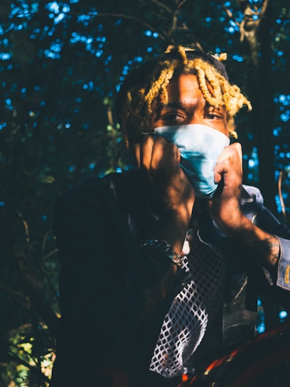 A person with blond curly hair is partially hiding their face with a blue surgical mask while sitting outdoors. Their hands are touching their face, and they are surrounded by foliage. The lighting creates dramatic shadows across their face and body.