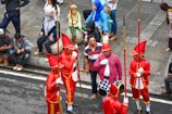 A group of performers dressed in bright red uniforms with tall hats and golden accents are holding long staffs, standing together on a street. They are surrounded by several onlookers, some of whom are sitting on a curb while others stand nearby. The onlookers are casually dressed, adding contrast to the performers' vibrant attire.