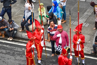 A group of performers dressed in bright red uniforms with tall hats and golden accents are holding long staffs, standing together on a street. They are surrounded by several onlookers, some of whom are sitting on a curb while others stand nearby. The onlookers are casually dressed, adding contrast to the performers' vibrant attire.
