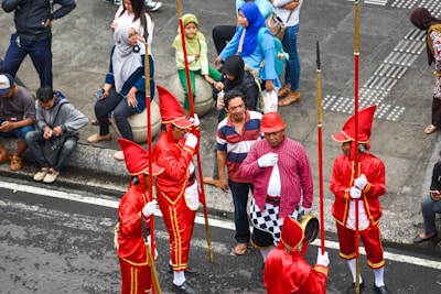 A group of performers dressed in bright red uniforms with tall hats and golden accents are holding long staffs, standing together on a street. They are surrounded by several onlookers, some of whom are sitting on a curb while others stand nearby. The onlookers are casually dressed, adding contrast to the performers' vibrant attire.