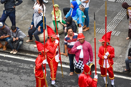 A group of performers dressed in bright red uniforms with tall hats and golden accents are holding long staffs, standing together on a street. They are surrounded by several onlookers, some of whom are sitting on a curb while others stand nearby. The onlookers are casually dressed, adding contrast to the performers' vibrant attire.