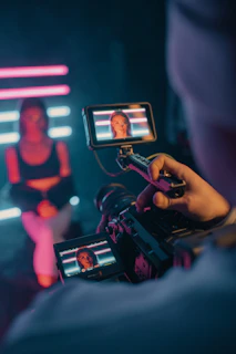 Close-up of a video camera filming a person sitting in front of neon lights. The person is blurred out in the background while their face is clearly visible on the camera's screen.