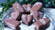 A collection of chocolate-covered desserts arranged on a silver tray. Some treats are in geometric heart shapes, while others resemble ice cream bars on wooden sticks. They are decorated with drizzles of chocolate and sprinkled with small pink and white candies. Green leaves add contrast to the overall presentation.