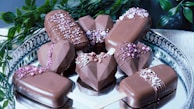 A collection of chocolate-covered desserts arranged on a silver tray. Some treats are in geometric heart shapes, while others resemble ice cream bars on wooden sticks. They are decorated with drizzles of chocolate and sprinkled with small pink and white candies. Green leaves add contrast to the overall presentation.
