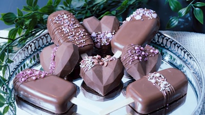 A collection of chocolate-covered desserts arranged on a silver tray. Some treats are in geometric heart shapes, while others resemble ice cream bars on wooden sticks. They are decorated with drizzles of chocolate and sprinkled with small pink and white candies. Green leaves add contrast to the overall presentation.