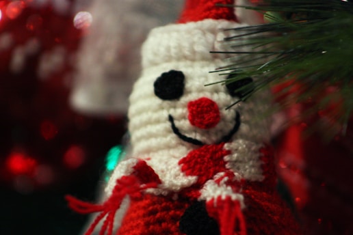 A crocheted snowman ornament featuring a red and white color scheme with black details for the eyes and buttons. Its cheerful smile is highlighted by a bright red nose. The background includes blurred festive decorations and green pine needles.