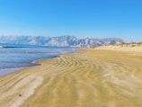 Expansive sandy beach with gentle waves lapping against the shore. Rugged cliffs create a dramatic backdrop under a clear blue sky.