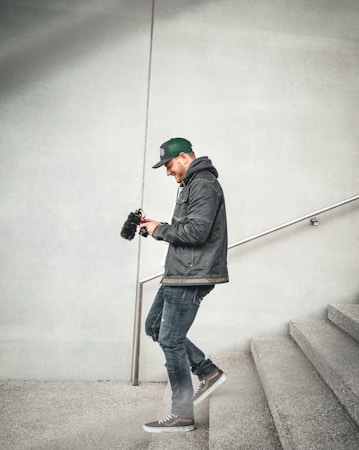 A person in a casual outfit is walking down a concrete staircase while looking at a camera with a microphone attached. The individual is wearing a dark jacket, jeans, and a green cap. The setting is minimalistic with a focus on the gray textures of the wall and stairs.