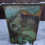 An old, rusted metal container with a weathered, turquoise paint surface stands in a snowy area. The metal shows signs of wear and corrosion, displaying layered patterns of rust in various shades of green, brown, and orange. The background is a fence, partially covered in snow, and there are sparse, dry plants on top of the container.