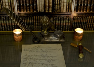 A desk is adorned with a decorative quill and ink set, featuring a lion sculpture. Two lit candles in copper holders provide ambient light. A vintage document with cursive handwriting lies on the table, surrounded by a wax seal and a key. Decorative bookshelves serve as the backdrop.