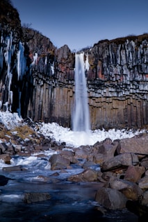 A stunning waterfall cascades down onto icy rocks surrounded by a wall of dark basalt columns. Above the waterfall, the sky is clear and blue. The area is enveloped in snow and ice, creating a serene winter landscape.
