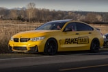 A yellow sports sedan is parked on the side of a road in a rural area, with a logo on the side that says 'FAKE TAXI'. The car has a sleek design with dark-tinted windows and black rims. In the background, there is a field with dry grass and leafless trees, and a mountain range is visible in the distance.
