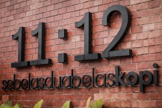 Large black numbers '11:12' are mounted on a red brick wall. Below the numbers, the words 'sebelasduabelaskopi' are displayed in bold black text.
