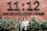 A person stands in front of a brick wall with large black text that reads '11:12' and additional smaller text spelling 'sebelas duabelas kopi'. Green foliage partially covers the lower portion of the wall.