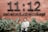 A person stands in front of a brick wall with large black text that reads '11:12' and additional smaller text spelling 'sebelas duabelas kopi'. Green foliage partially covers the lower portion of the wall.