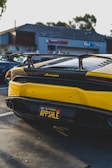 A yellow Lamborghini with a custom license plate is parked in a parking lot with a building in the background. The car has a prominent rear spoiler and glossy finish. Other vehicles are visible in the lot, and there are trees and a clear sky.