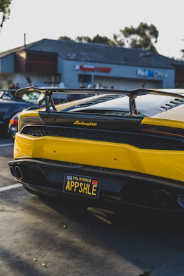 A yellow Lamborghini with a custom license plate is parked in a parking lot with a building in the background. The car has a prominent rear spoiler and glossy finish. Other vehicles are visible in the lot, and there are trees and a clear sky.
