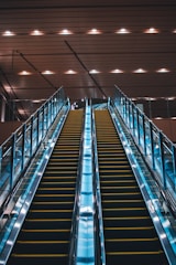 Twin escalators ascend in parallel under a ceiling with evenly spaced lights. The escalators are enclosed in glass with metal railings and have a sleek, modern appearance. The lighting creates a reflective effect on the shiny surfaces.
