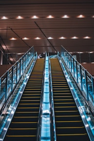 Twin escalators ascend in parallel under a ceiling with evenly spaced lights. The escalators are enclosed in glass with metal railings and have a sleek, modern appearance. The lighting creates a reflective effect on the shiny surfaces.